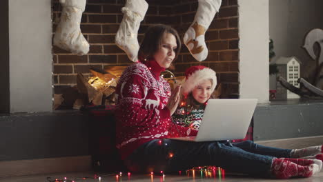 Glückliche-Mutter-Und-Tochter-In-Weihnachtspullovern-Und-Mit-Lichtern-Um-Den-Hals,-Die-Auf-Dem-Boden-Sitzen,-Während-Sie-Einen-Laptop-Benutzen