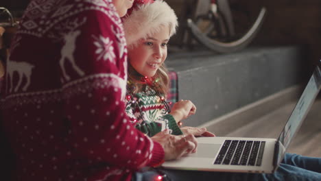 Glückliches-Kleines-Mädchen-In-Weihnachtspullover-Und-Weihnachtsmütze-Sitzt-Mit-Ihrer-Mutter-Auf-Dem-Boden-Und-Schaut-Etwas-Auf-Laptop-computer