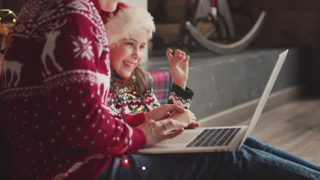 Glückliches-Kleines-Mädchen-In-Weihnachtspullover-Und-Weihnachtsmütze-Sitzt-Mit-Ihrer-Mutter-Auf-Dem-Boden-Und-Schaut-Etwas-Auf-Laptop-1