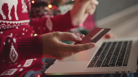 Close-Up-Of-An-Unrecognizable-Woman-In-Christmas-Sweater-Holding-Credit-Card-And-Shopping-Online-With-Laptop-Computer