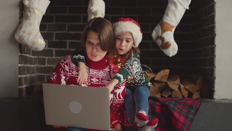 Glückliche-Mutter-Und-Tochter-In-Weihnachtspullovern-Und-Mit-Lichtern-Um-Den-Hals-Sitzen-Auf-Dem-Sofa,-Während-Sie-Einen-Laptop-Benutzen
