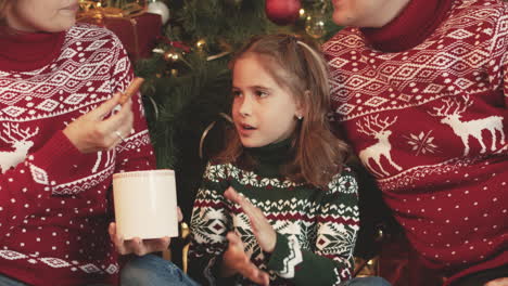 Glückliche-Familie-In-Weihnachtspullovern,-Die-Zu-Hause-Vor-Dem-Weihnachtsbaum-Zusammensitzen,-Während-Die-Mutter-Das-Kleine-Mädchen-Mit-Einem-Keks-Füttert