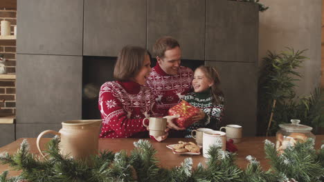 Liebevolle-Eltern-Geben-Ihrer-Kleinen-Tochter-Ein-überraschungsgeschenk,-Während-Sie-Am-Weihnachtstag-Zu-Hause-Zusammen-Mit-Heißen-Getränken-Und-Keksen-Frühstücken