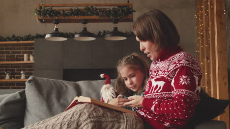 Schöne-Mutter-Sitzt-Mit-Entzückender-Tochter-Auf-Der-Couch-Und-Liest-Ihr-Märchen-Am-Weihnachtstag-Zu-Hause-3