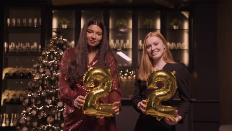 Zwei-Freundinnen-In-Eleganter-Kleidung,-Die-Luftballons-Mit-Den-Nummern-22-Halten,-Während-Sie-Auf-Der-Neujahrsparty-Tanzen