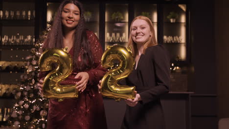 Two-Women-Friends-Wearing-Elegant-Clothes-Holding-Balloons-With-The-Numbers-22-While-Dancing-In-New-Year's-Party-1