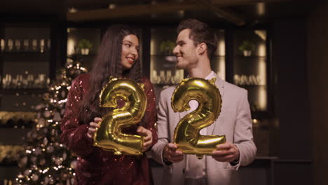 Woman-And-Man-Wearing-Elegant-Clothes-Holding-Balloons-With-The-Numbers-22-Kiss-Each-Other-While-Dancing-In-New-Year's-Party