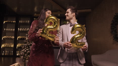 Frau-Und-Mann-In-Eleganter-Kleidung,-Die-Luftballons-Mit-Den-Zahlen-22-Halten,-Während-Sie-Tanzen-Und-Sich-Auf-Der-Neujahrsparty-Küssen