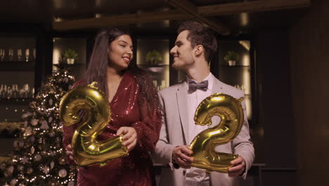 Woman-And-Man-Wearing-Elegant-Clothes-Holding-Balloons-With-The-Numbers-22-While-Dancing,-Kissing-And-Hugging-Each-Other-In-New-Year's-Party
