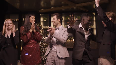 Group-Of-Friends-Wearing-Elegant-Clothes-Blow-And-Throw-Conffeti-While-Dancing-And-Hugging-Each-Other-In-New-Year's-Party