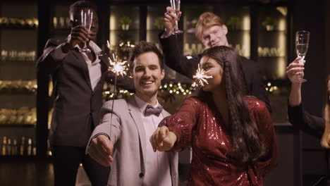 Camera-Focuses-On-A-Couple-Celebrating-New-Year-Eve-While-Holding-Sparkles-And-Kiss-Each-Other,-Their-Friends-Are-Behind-Raising-Glasses-Of-Champagne