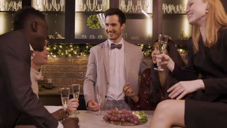 Group-Of-Friends-Dressed-In-Elegant-Clothes-Celebrating-The-New-Year's-Party,-They-Stand-Around-The-Bar-Counter-While-Drinking-Champagne-And-Eating-Grapes