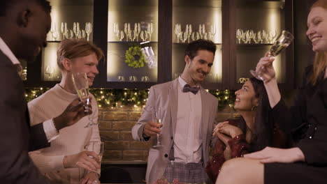 Group-Of-Friends-Dressed-In-Elegant-Clothes-Celebrating-The-New-Year's-Party,-They-Stand-Around-The-Bar-Counter-While-Toasting-And-Drinking-Champagne