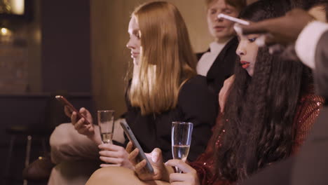 Group-Of-Friends-Dressed-In-Stylish-Clothes-Sitting-On-The-Sofa-While-Using-Smartphones-And-Drinking-Champagne-During-New-Year-Eve-Party