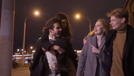 Two-Couples-Of-Friends-Dressed-In-Elegant-Clothes-Walking-In-The-Street-Street-While-Talking-And-Laughing-After-New-Year's-Party,-One-Of-The-Girls-Is-Perched-On-Her-Boyfriend's-Back