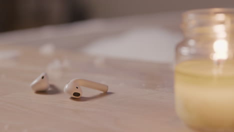 Close-Up-View-Of-Earphones-And-A-Candle-On-Top-Of-A-Wooden-Table,-In-The-Background-A-Blurred-Bathtub-Filled-With-Water-And-Foam