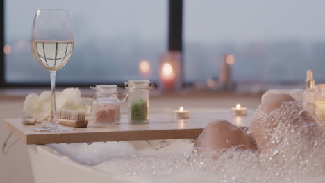 Camera-Focuses-On-A-Wooden-Board-Above-A-Bathtub-With-A-Glass-Of-Wine,-Bath-Salts-And-A-Candle