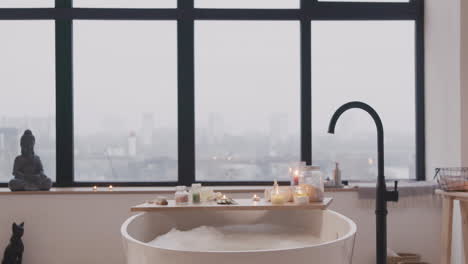 Die-Kamera-Fokussiert-Eine-Weiße-Badewanne-Mit-Wasser-Und-Schaum,-Einen-Holztisch-Mit-Badezimmerelementen-Und-Ein-Großes-Fenster-Im-Hintergrund