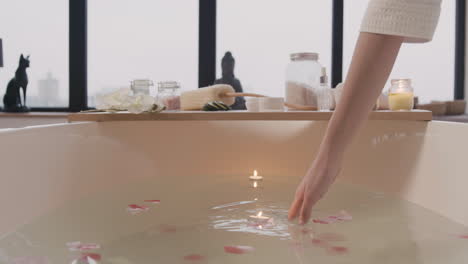 Die-Kamera-Konzentriert-Sich-Auf-Eine-Weiße-Badewanne-Mit-Wasser,-Blumen-Und-Kerzen-Und-Einen-Holztisch-Mit-Badezimmerelementen