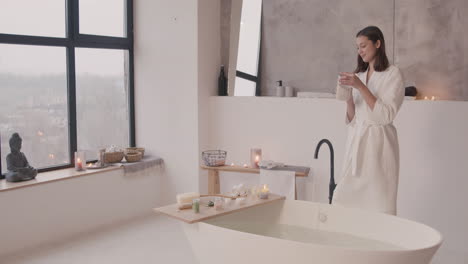 Woman-In-Bathrobe-Pours-Bath-Salts-Into-The-Bathtub-And-Then-Touches-The-Water
