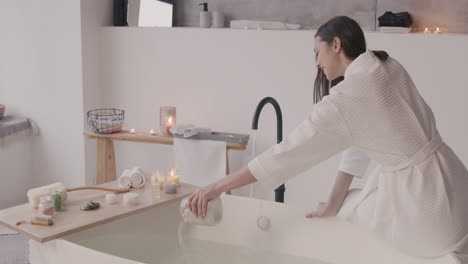 Woman-In-Bathrobe-Sitting-On-Edge-Of-Bathtub-While-Pouring-Bath-Salts-On-The-Water