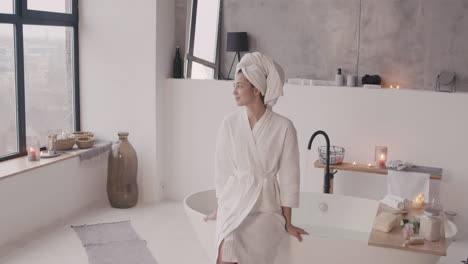 Front-View-Of-A-Woman-Sitting-On-The-Edge-Of-Bathtub-Posing-And-Smiling-At-The-Camera-With-A-Towel-On-Her-Head-And-A-Bathrobe