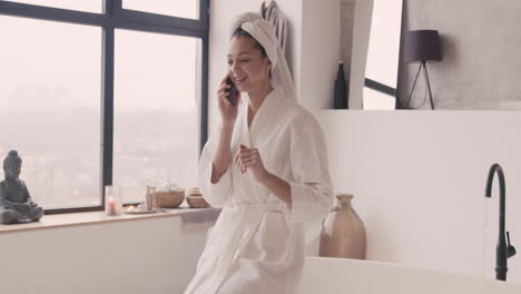 Woman-In-Bathrope-With-A-Towel-In-Her-Head-Talking-On-The-Phone-Sitting-On-The-Edge-Of-Bathtub