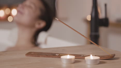 Close-Up-View-Of-A-Table-With-Candles-And-Incense,-In-The-Background-A-Blurred-Woman-Taking-A-Bath-And-Drinking