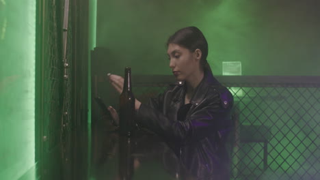 Serious-Girl-In-Leather-Jacket-Drinking-Beer-And-Using-Mobile-Phone-While-Sitting-On-A-Stool-At-Disco