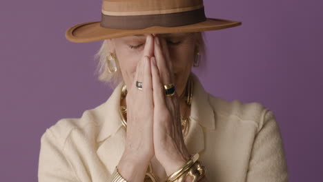 Blonde-Mature-Woman-With-Blue-Eyes-Dressed-In-Jacket,-Hat-And-Accessories-Posing-With-Hands-On-Her-Face-And-Smiling-At-Camera-On-Purple-Background