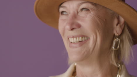 Close-Up-View-Of-Blonde-Mature-Woman-With-Blue-Eyes-Dressed-In-Jacket-And-Hat-Posing-Wiping-Tears-And-Smiling-At-Camera-On-Purple-Background
