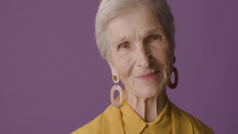 Senior-Woman-With-Short-Hair-Wearing-Mustard-Colored-Shirt-And-Jacket-And-Earrings-Posing-And-Smiling-At-Camera-On-Purple-Background
