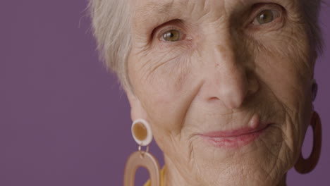 Close-Up-View-Of-Senior-Woman-With-Short-Hair-And-Green-Eyes-Wearing-Earrings-Posing-And-Smiling-At-Camera-On-Purple-Background