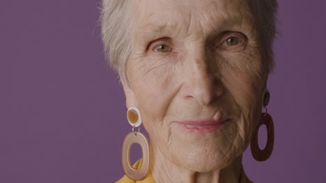 Close-Up-View-Of-Senior-Woman-Face-With-Short-Hair-And-Green-Eyes-Wearing-Earrings-Posing-And-Smiling-At-Camera-On-Purple-Background