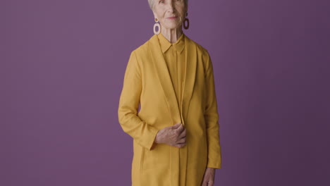 Senior-Woman-With-Short-Hair-Wearing-Mustard-Colored-Shirt-And-Jacket-And-Earrings-Posing-And-Smiling-At-Camera-On-Purple-Background-1