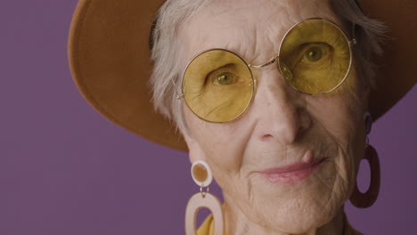 Nahaufnahme-Einer-älteren-Frau-Mit-Kurzen-Haaren,-Die-Hut,-Sonnenbrille-Und-Ohrringe-Trägt-Und-Auf-Violettem-Hintergrund-Posiert-Und-In-Die-Kamera-Blickt