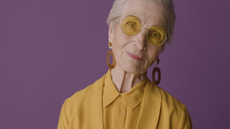 Seniorin-Mit-Kurzen-Haaren,-Senffarbenem-Hemd-Und-Jacke,-Ohrringen-Und-Sonnenbrille,-Posiert-Und-Blickt-In-Die-Kamera-Auf-Violettem-Hintergrund
