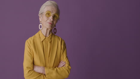 Senior-Woman-With-Short-Hair-Wearing-Mustard-Colored-Shirt-And-Jacket,-Earrings-And-Sunglasses-Posing-With-Crossed-Arms-And-Looking-At-Camera-On-Purple-Background
