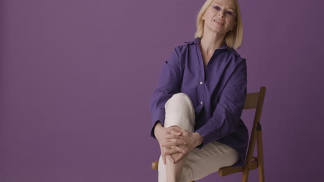 Blonde-Mature-Woman-With-Purple-Shirt,-Beige-Pants-And-Sunglasses-Posing-Looking-At-Camera-Sitting-On-Chair-With-Crossed-Legs-On-Purple-Background