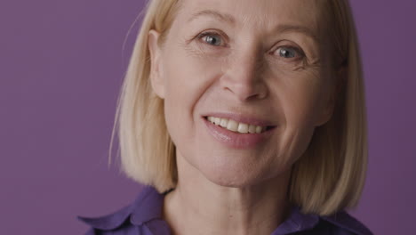 Close-Up-View-Of-Blonde-Mature-Woman-With-Green-Eyes-Smiling-And-Looking-At-Camera-On-Purple-Background