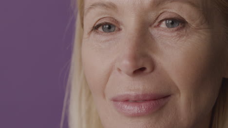 Close-Up-View-Of-Blonde-Mature-Woman-With-Green-Eyes-Smiling-And-Looking-At-Camera-On-Purple-Background-1