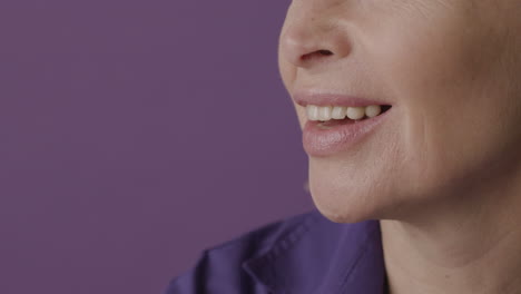 Close-Up-View-Of-Half-Face-Of-Blonde-Mature-Woman-With-Purple-Shirt-Smiling-On-Purple-Background