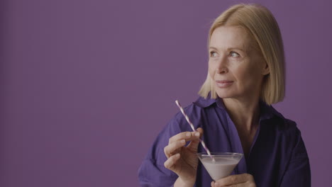 Blonde-Mature-Woman-With-Purple-Shirt-Posing-Holding-A-Cocktail-And-Smiling-At-Camera-On-Purple-Background