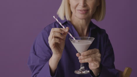 Blonde-Reife-Frau-Mit-Lila-Hemd-Posiert-Mit-Einem-Cocktail-Und-Lächelt-In-Die-Kamera-Auf-Lila-Hintergrund-1