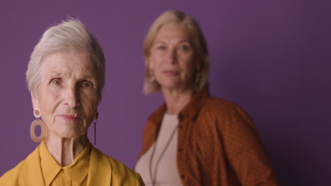 Blonde-Senior-Woman-With-Short-Hair-Wearing-Mustard-Colored-Shirt-And-Jacket-And-Earrings,-Posing-With-Blurred-Mature-Woman-On-Purple-Background