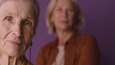 Close-Up-View-Of-Blonde-Senior-Woman-With-Short-Hair-Wearing-Mustard-Colored-Shirt-And-Jacket-And-Earrings,-Posing-With-Blurred-Mature-Woman-On-Purple-Background
