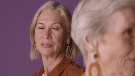 Nahaufnahme-Einer-Blonden-Reifen-Frau-Mit-Braunem-Hemd,-Die-Mit-Verschwommenen-älteren-Frauen-Mit-Kurzen-Haaren-Auf-Violettem-Hintergrund-Posiert