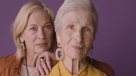 Close-Up-View-Of-Blonde-Mature-Woman-With-Brown-Shirt-Posing-With-Senior-Woman-With-Short-Hair-On-Purple-Background