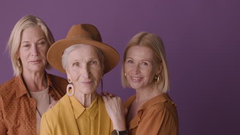 Seniorin-Mit-Hut-Und-Senfhemd-Posiert-Mit-Zwei-Blonden-Reifen-Frauen-Auf-Violettem-Hintergrund