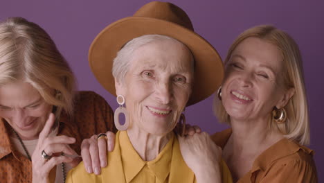 Seniorin-Mit-Hut-Und-Senfhemd-Posiert-Mit-Zwei-Blonden-Reifen-Frauen-Auf-Violettem-Hintergrund,-Die-In-Die-Kamera-Lächeln